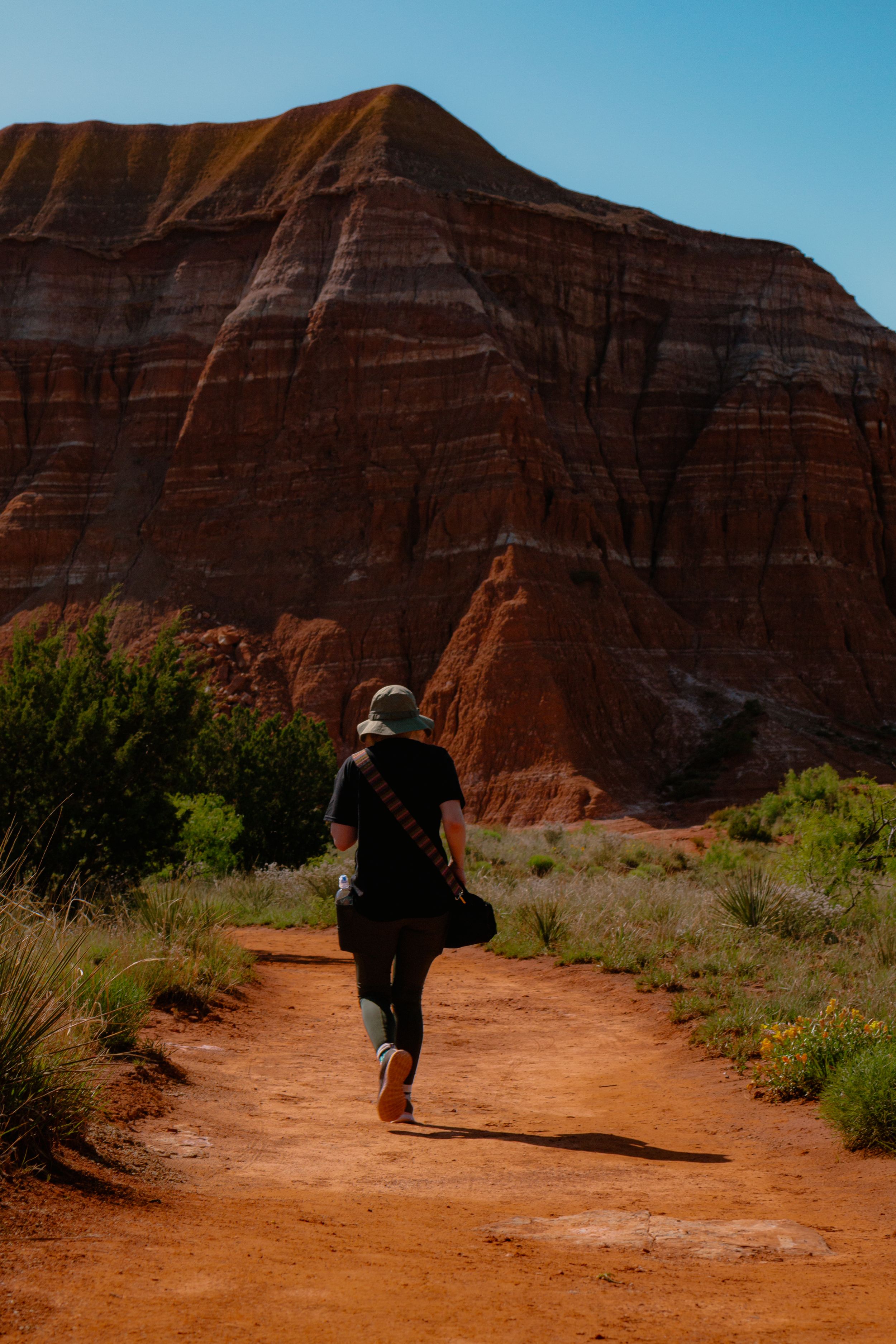 A hiking trail winds through red rock formations in a desert landscape with scrub vegetation and clear blue skies.