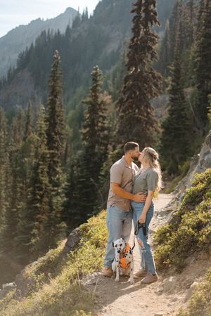 A couple and their Dalmatian dog share a tender moment on a mountain trail surrounded by evergreen trees.