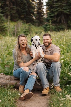 Couple and their dalmatian puppy share joyful moments while sitting on a fallen log in a lush mountain meadow.