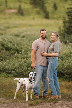 A couple in casual attire poses with their Dalmatian dog in a scenic mountain meadow.