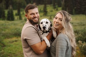 A couple and their dog share a happy moment together in a mountain setting with pine trees.