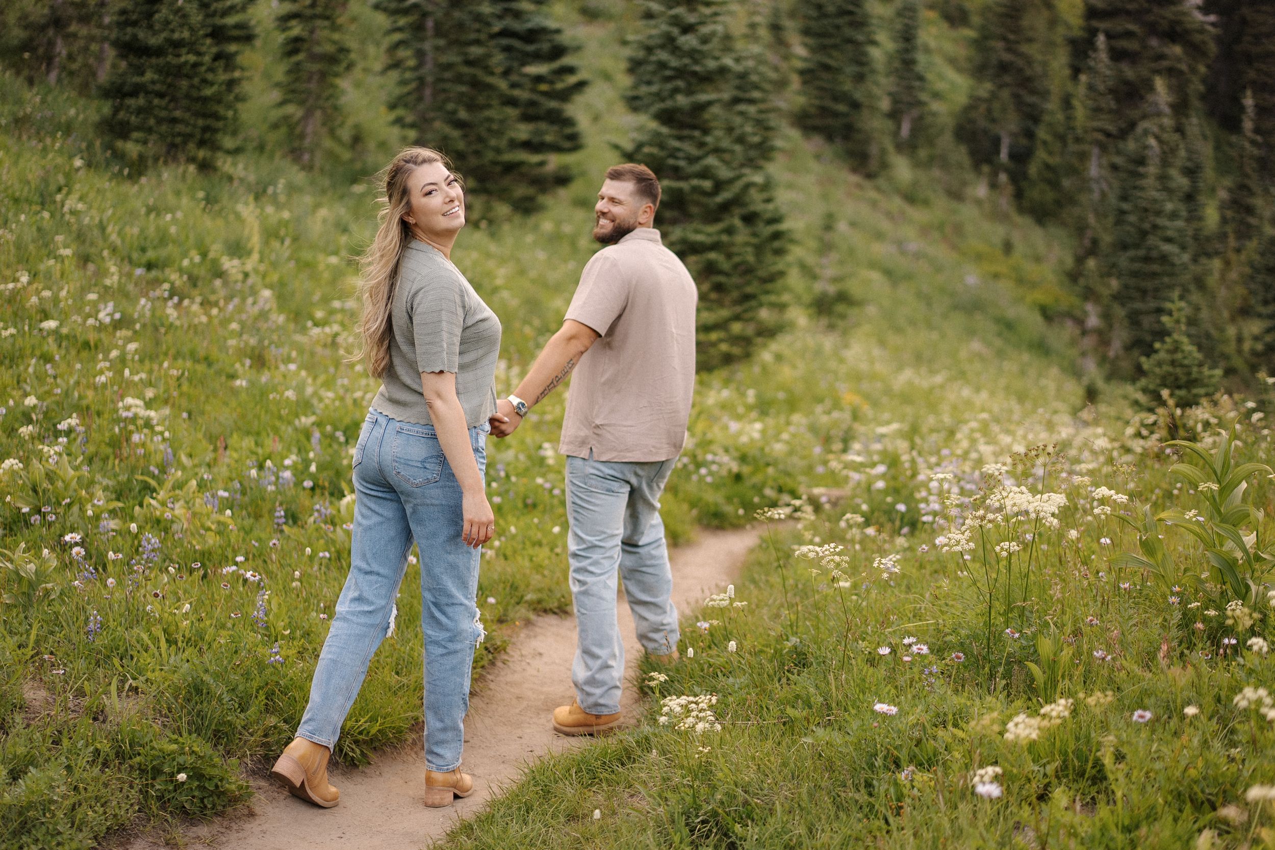 Two people walk hand in hand along a wildflower-filled mountain trail surrounded by evergreen trees.