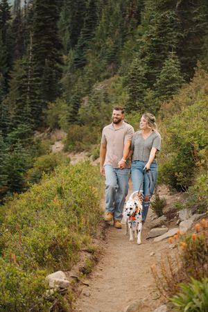 A couple walks their spotted dog along a scenic mountain hiking trail surrounded by evergreen trees and foliage.