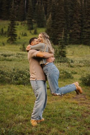 A couple embraces joyfully in a mountain meadow surrounded by evergreen trees on a sunny day.