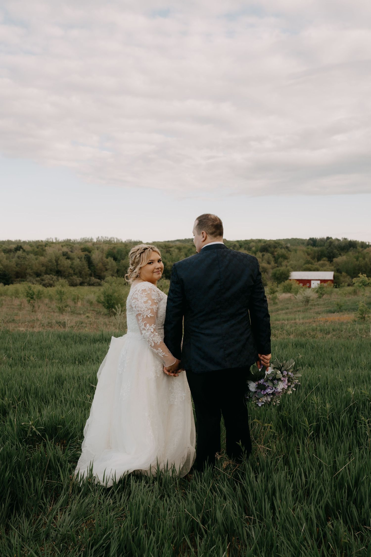 A couple in wedding attire walks through a lush green meadow on their special day.