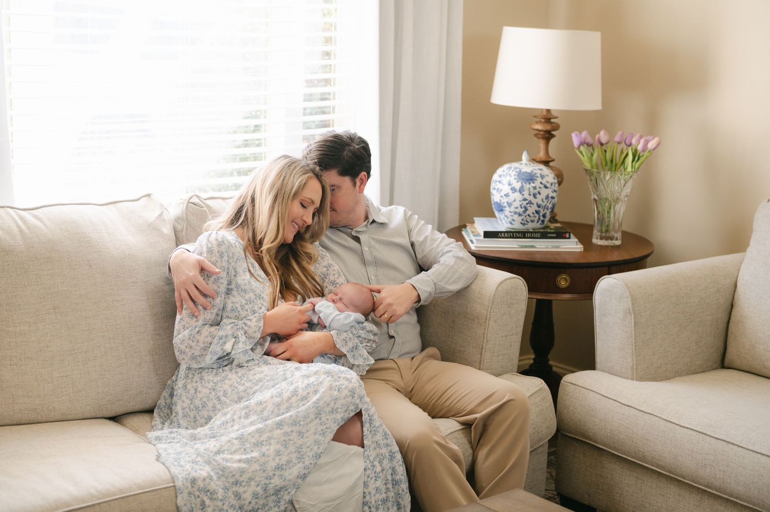 Couple sitting on cream sofa tenderly holding newborn baby in cozy home setting.