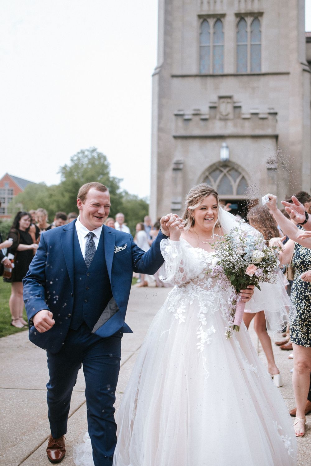 Newlyweds walk down church steps while guests throw confetti during joyful wedding exit celebration.