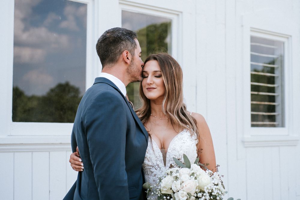Romantic outdoor wedding moment with couple in formal attire and white bouquet against white building exterior.