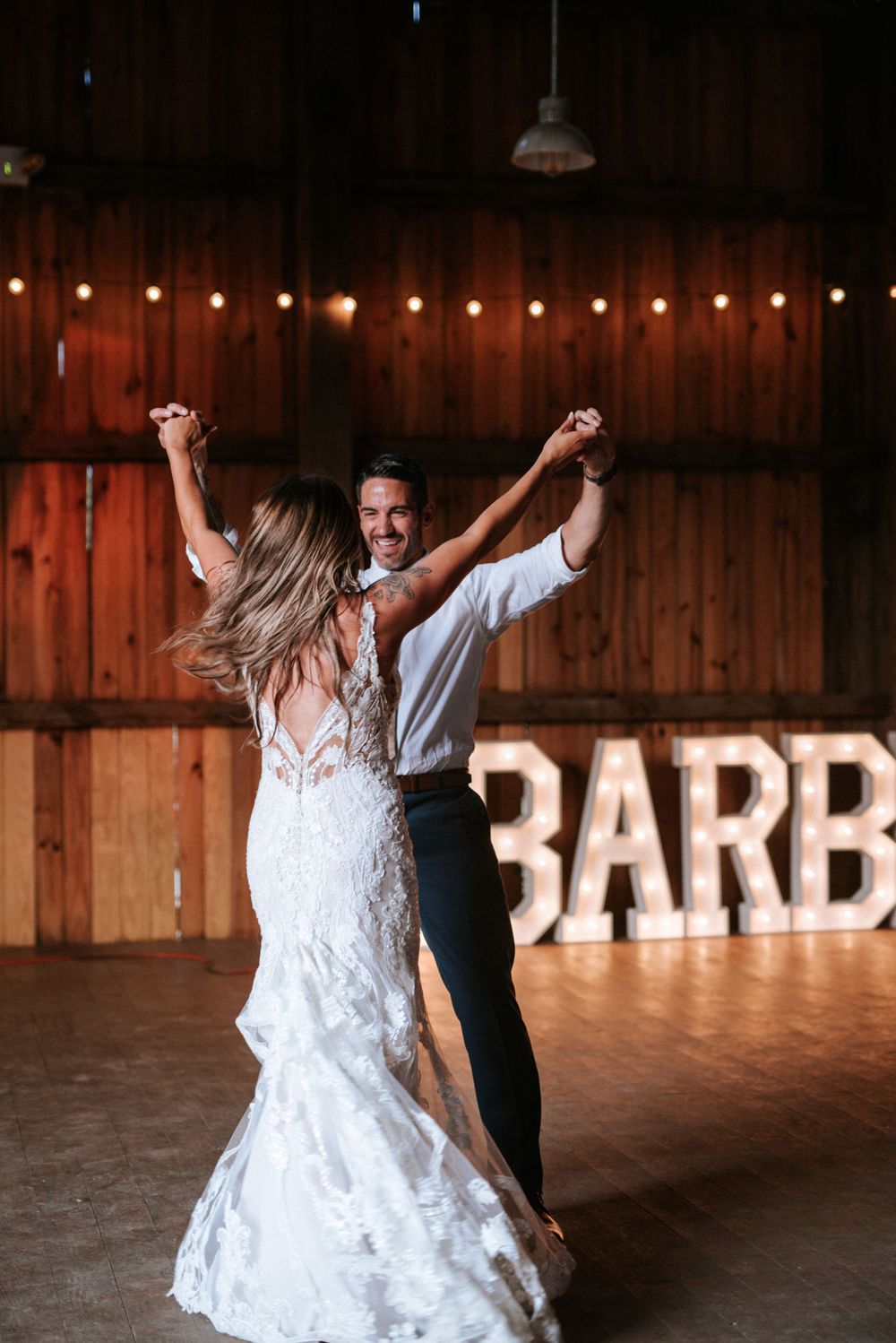 A couple dances joyfully in a rustic barn venue with illuminated 'BARN' sign in background.