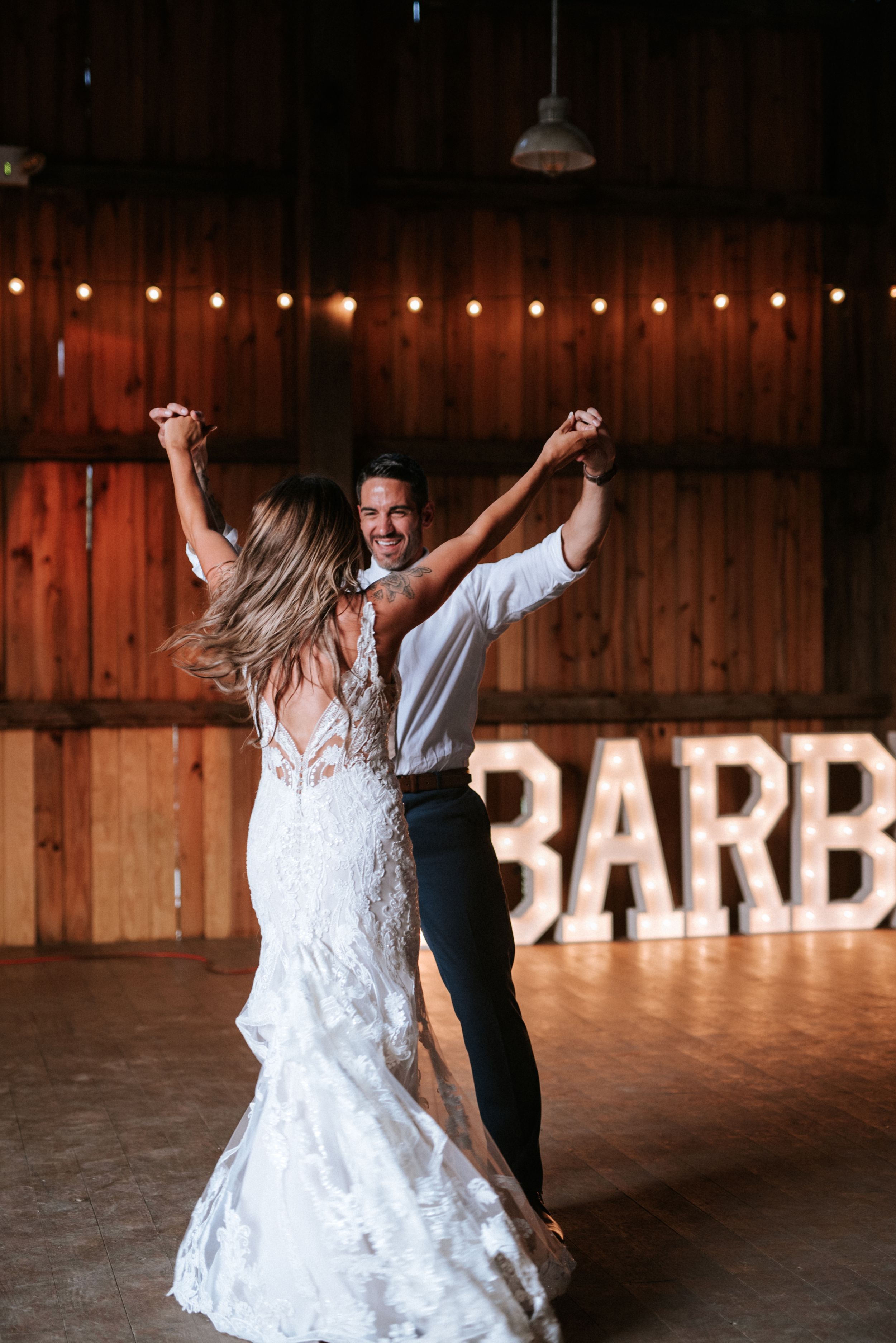 A couple dances joyfully in a rustic barn venue with illuminated 'BARN' sign in background.