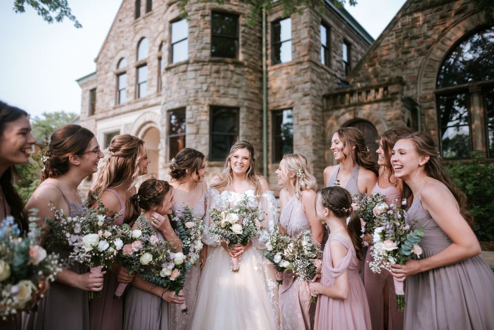 A bridal party in pink dresses poses with bouquets outside a stone building.