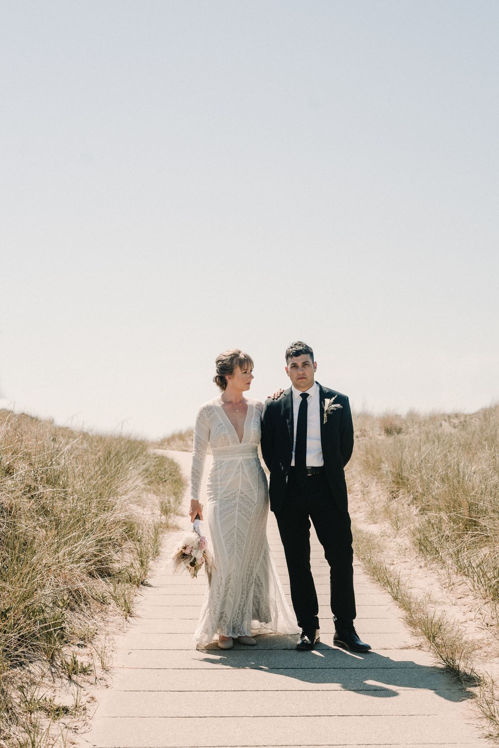 Beach boardwalk wedding portrait of couple in formal attire against sandy dunes.