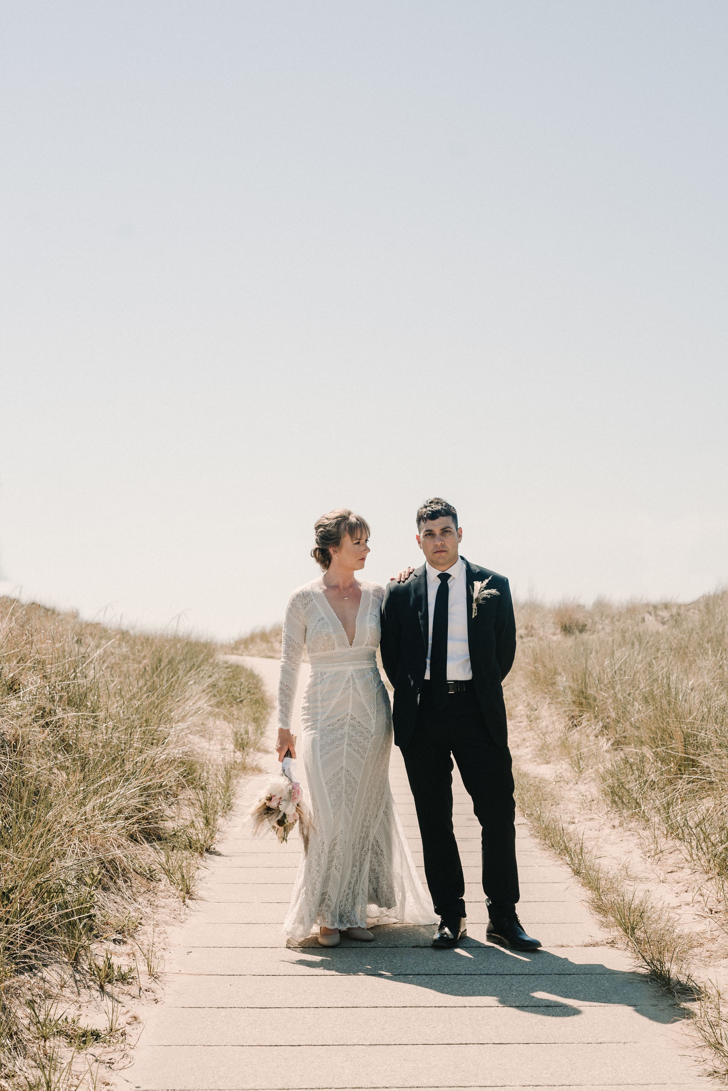 Beach boardwalk wedding portrait of couple in formal attire against sandy dunes.