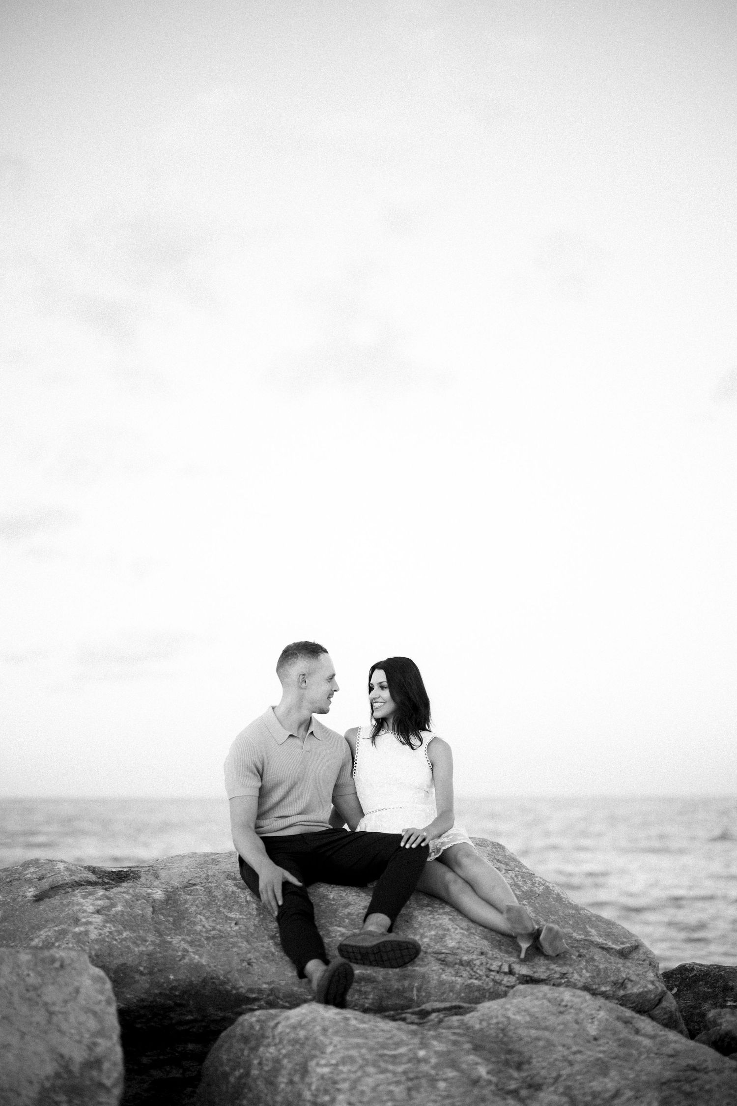 A romantic black and white photo of a couple sitting together on coastal rocks by the ocean.