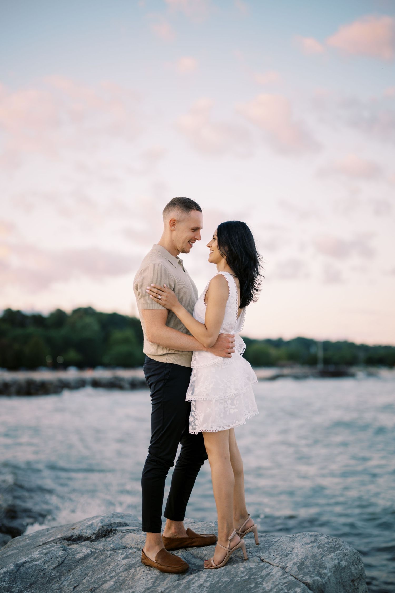 A romantic couple embraces on rocky shoreline at sunset with water and pink clouds in background.