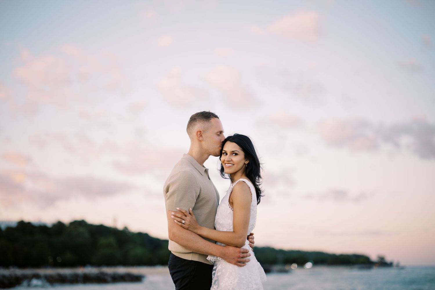 Couple embraces on beach at sunset with pink clouds and calm waters in background.