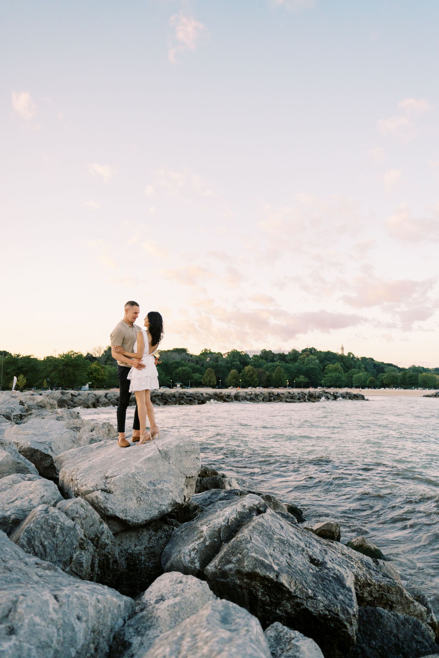 A couple stands together on rocky shoreline during sunset with water and trees in background.