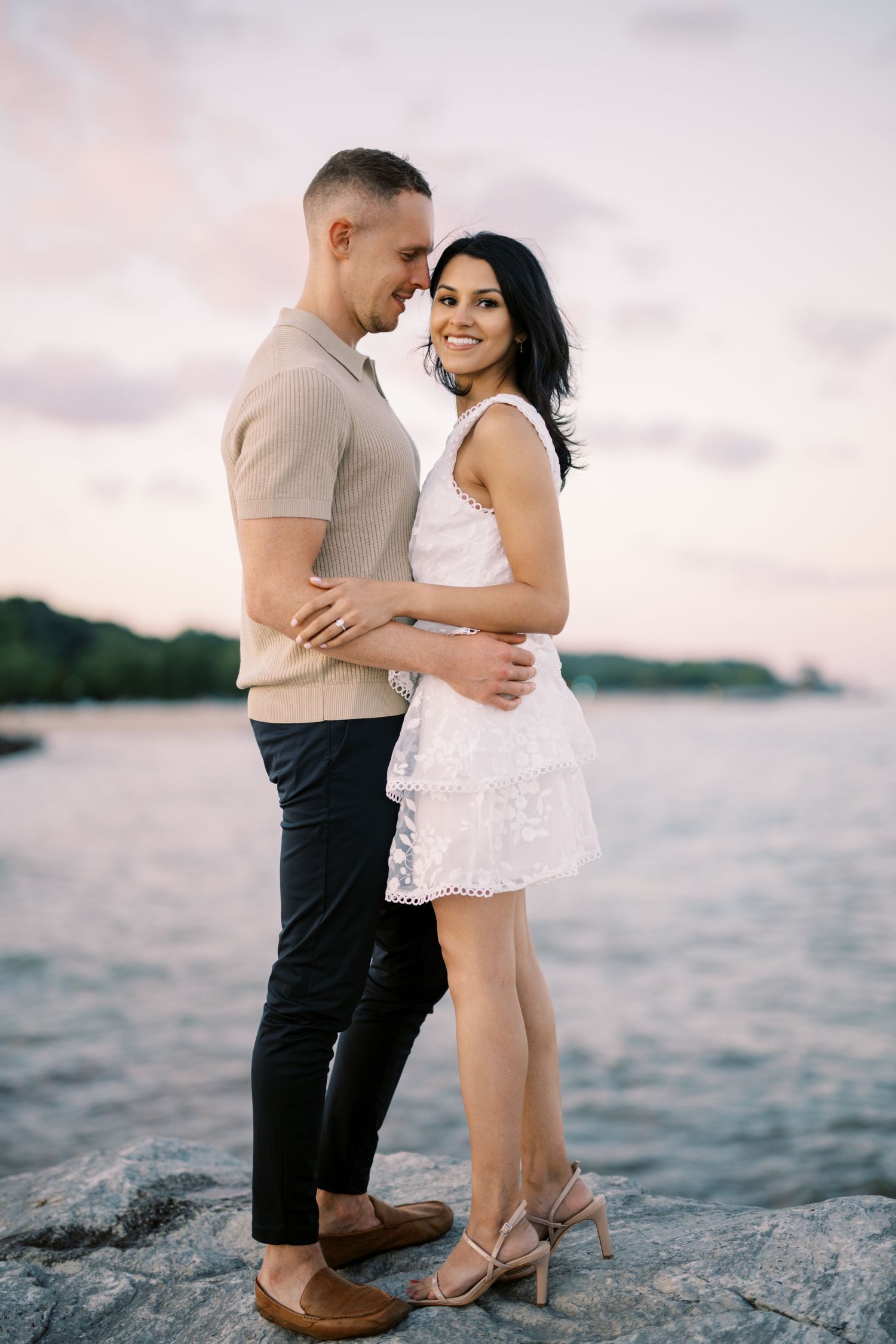 A romantic couple embraces on coastal rocks at sunset, with her in a white dress and him in casual attire.