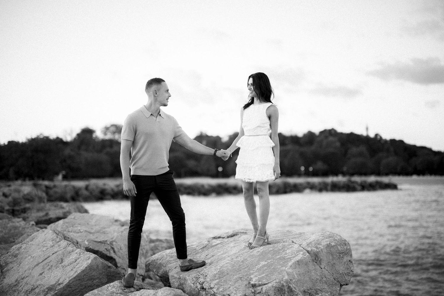 A couple in casual attire stands on rocky shoreline during sunset in black and white photo.