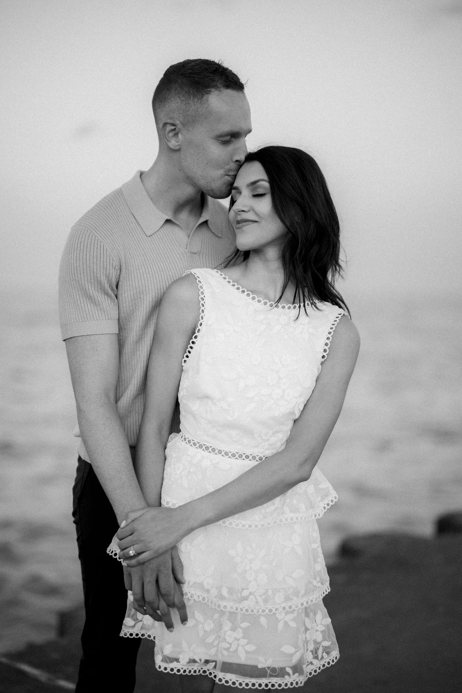 Black and white portrait of intimate couple in formal attire embracing near water.