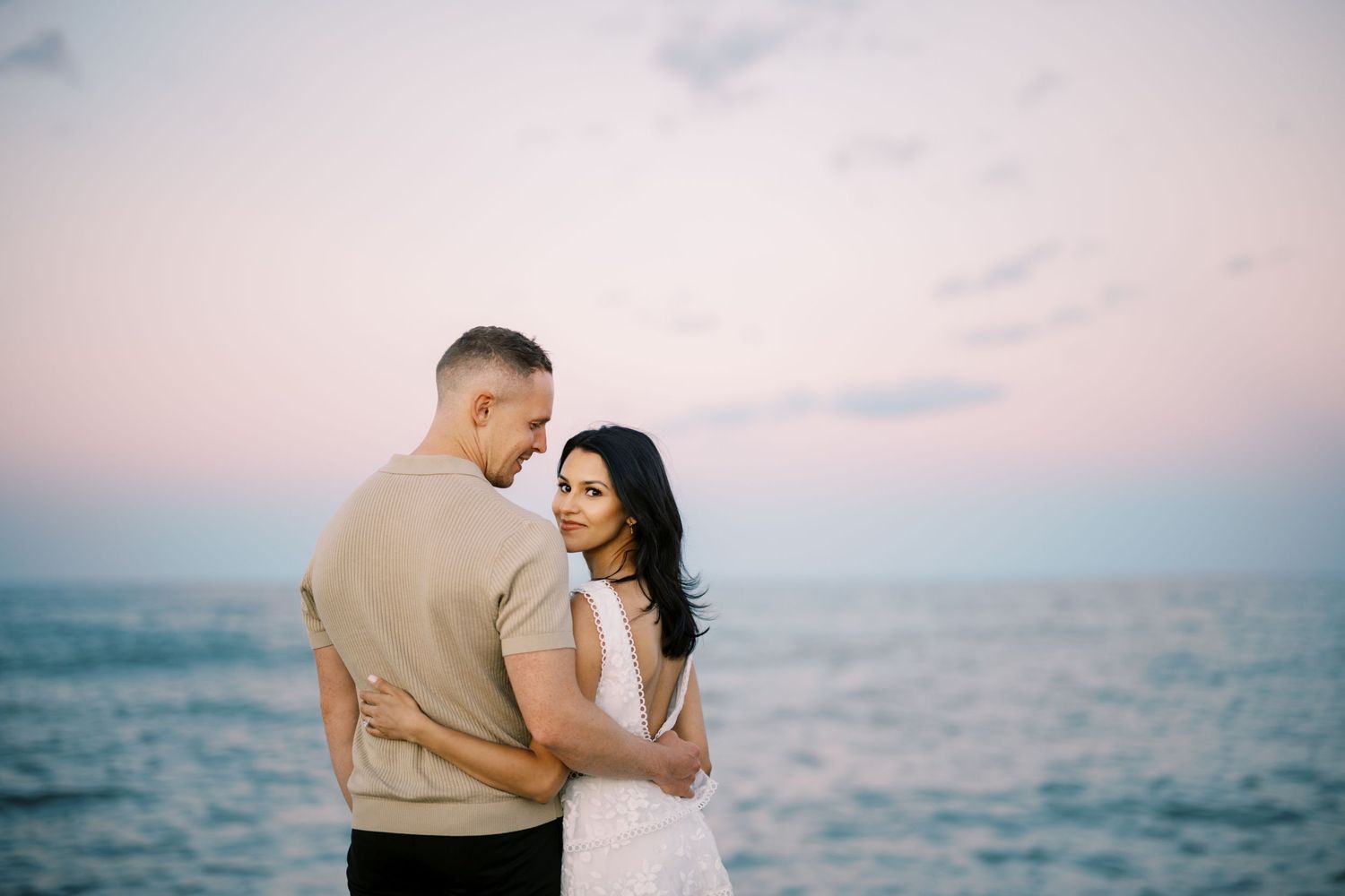 A romantic couple embraces near the ocean during sunset, with soft pink skies and calm waters in the background.