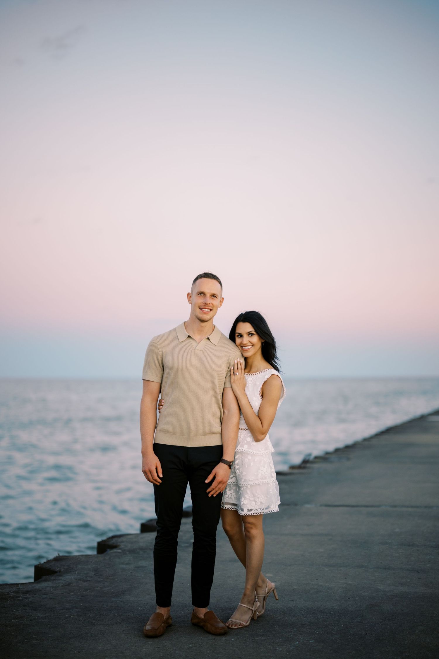 Couple poses together on pier at dusk with pink sky and ocean in background.