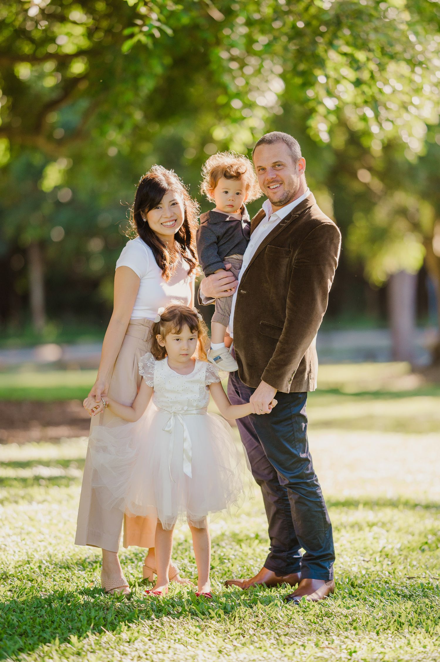 Family portrait outdoors in park with sunlight filtering through trees.