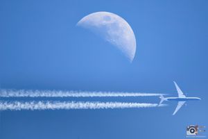 Passenger jet flies across bright blue sky leaving white contrails against fading crescent moon backdrop.