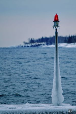 Ice-covered nautical marker with red beacon light against a winter seascape.