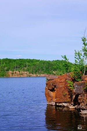 Rugged red rock cliffs along a serene blue lake shoreline with green forest in the background under clear skies.