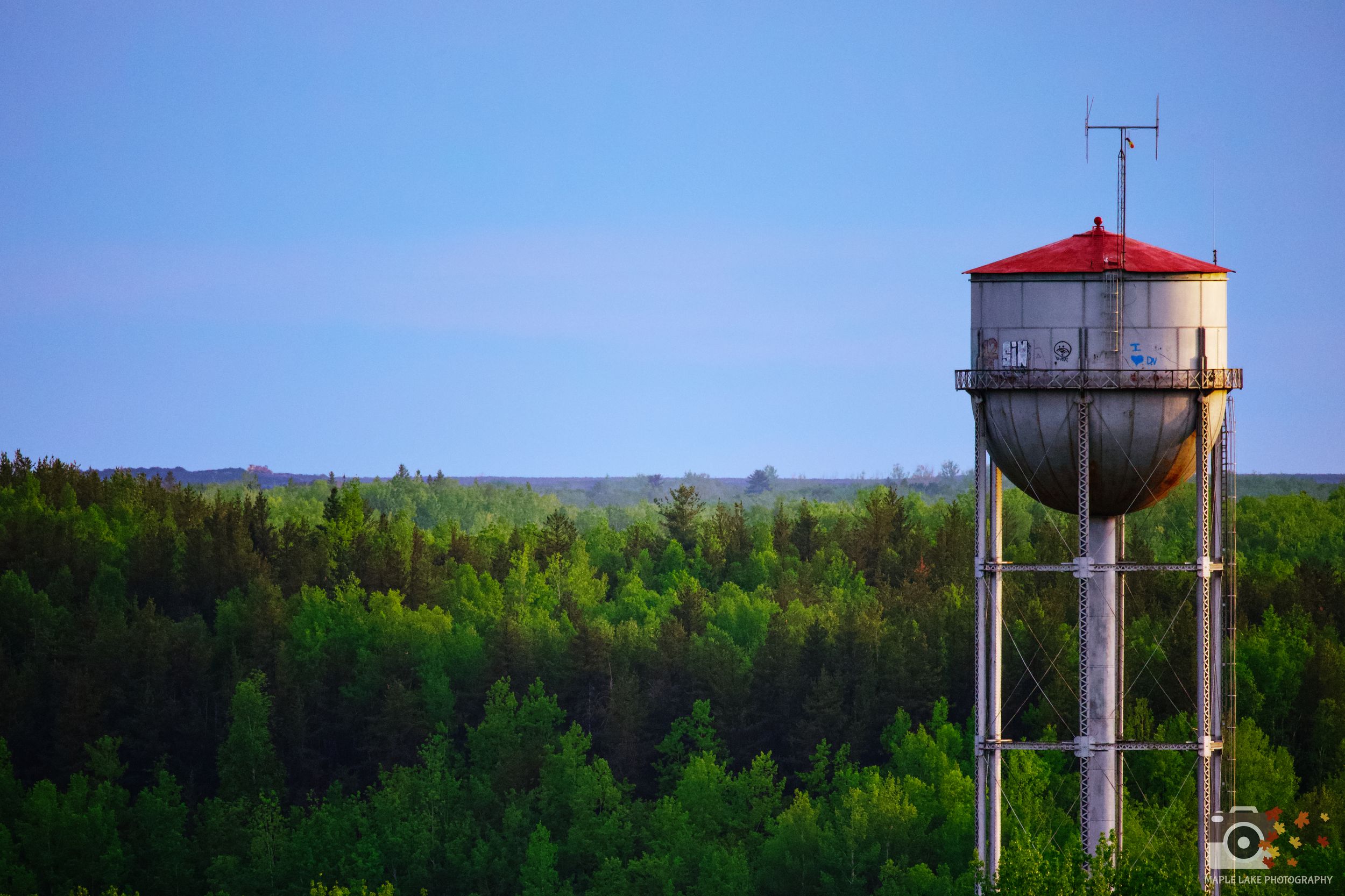 A tall metal water tower with a cylindrical tank and red roof stands prominently against a forest landscape at dusk.