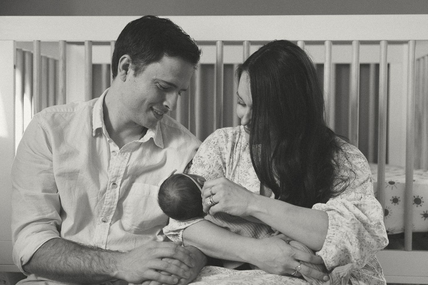 Parents sharing tender moment with newborn baby in white nursery, photographed in black and white.