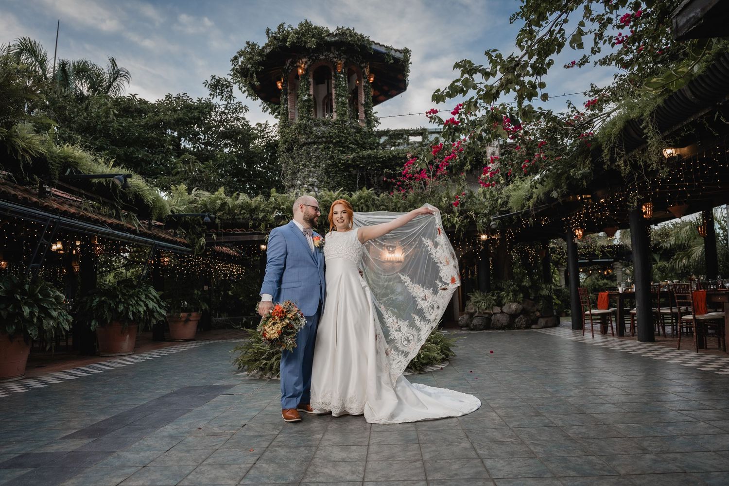 A couple embraces on a wet patio surrounded by lush greenery and pink flowers at an outdoor venue.