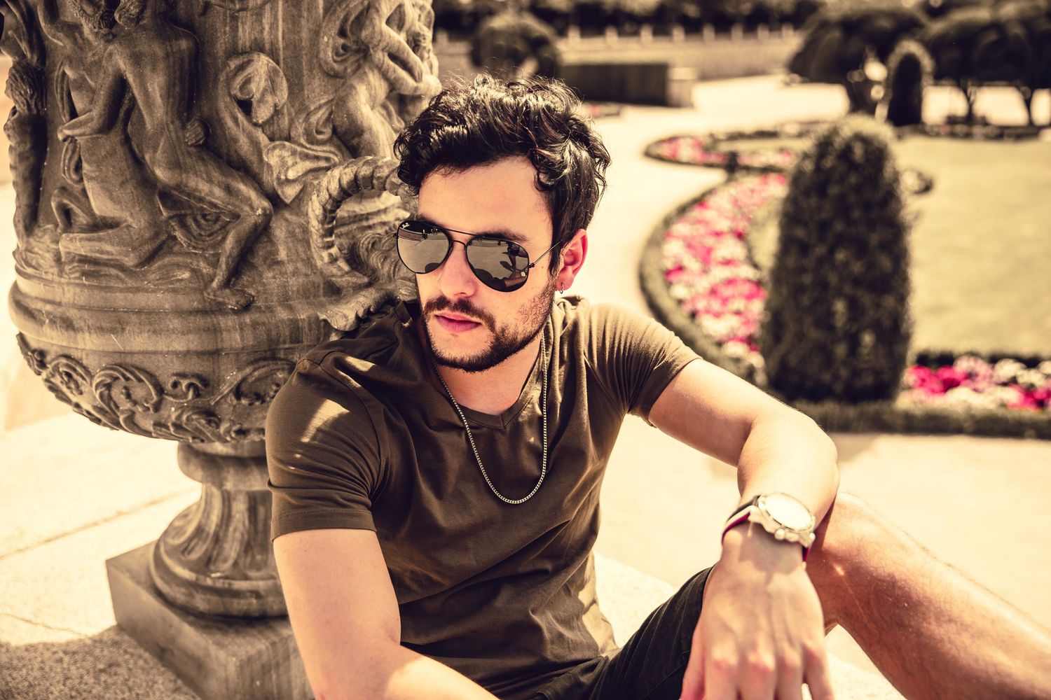 Person wearing sunglasses relaxing by ornate stone sculpture in a garden setting.