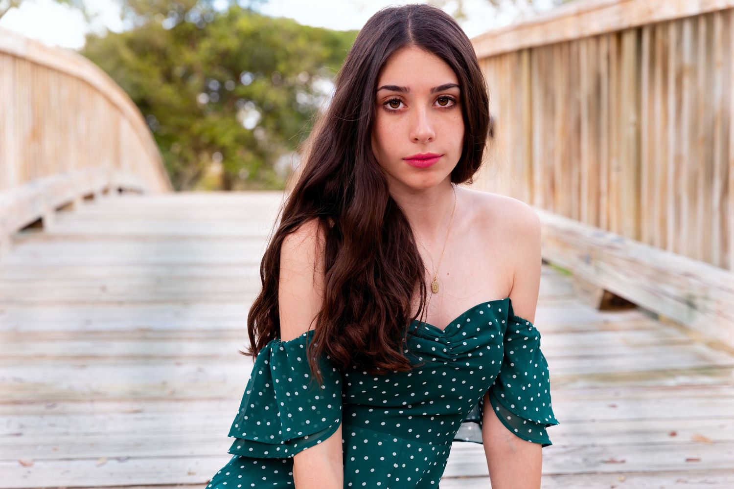 Woman in a green polka dot off-shoulder dress poses on a wooden bridge outdoors.