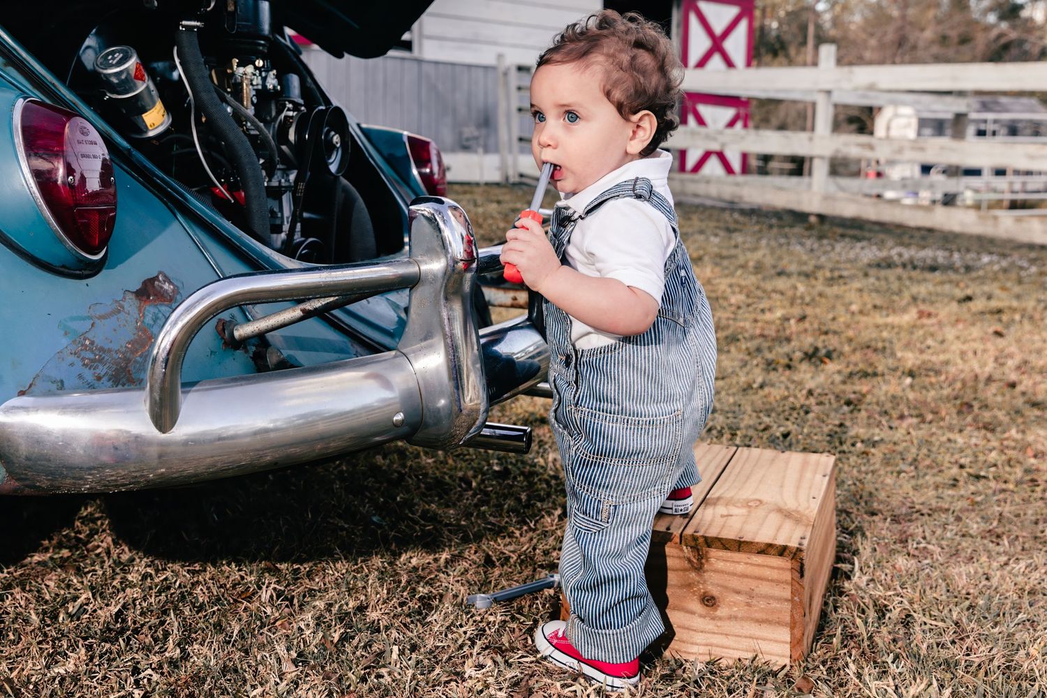 Small toddler in denim overalls standing on wooden crate near vintage motorcycle.