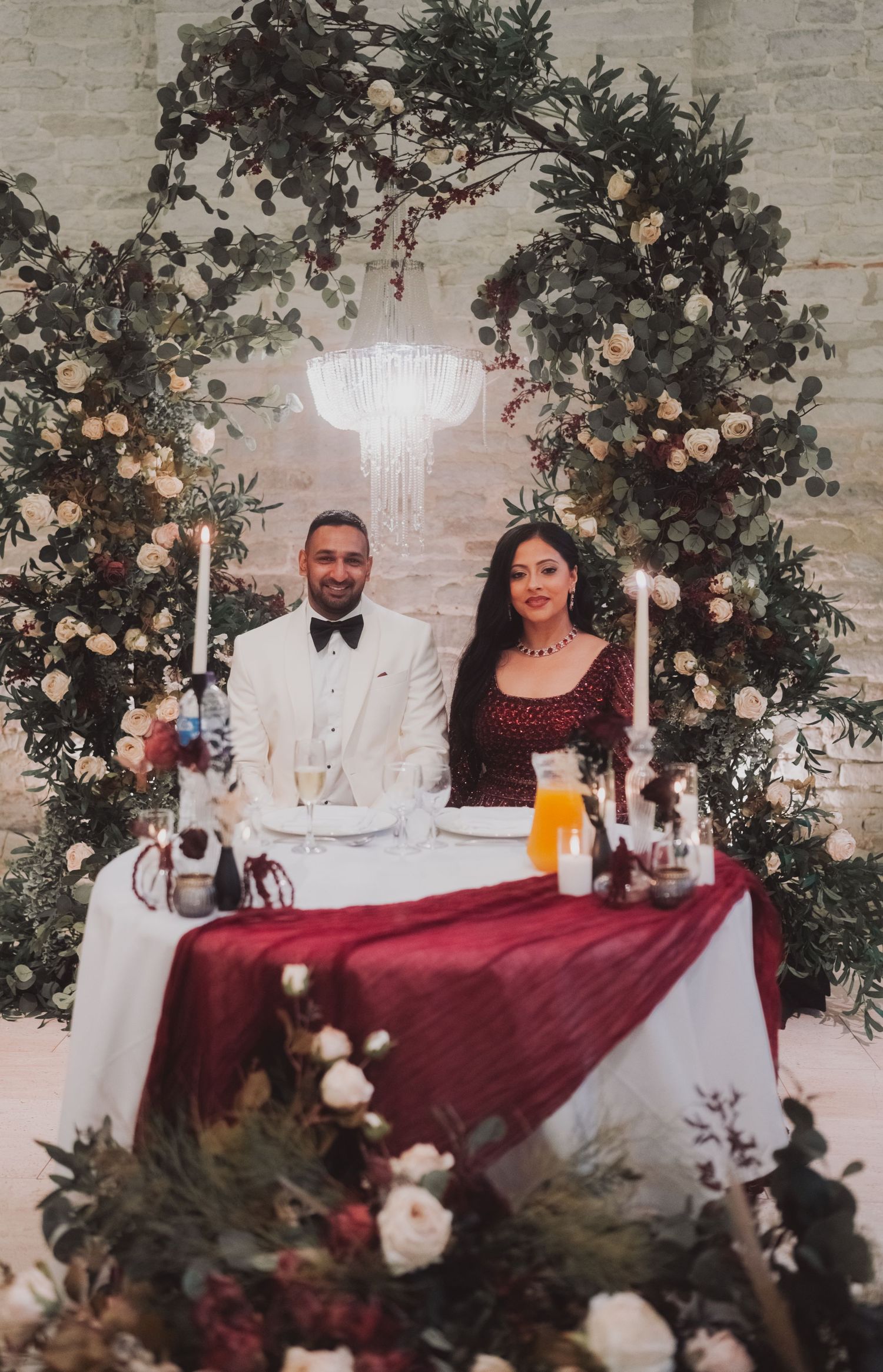 Elegant wedding reception setup with floral arch, chandelier, and burgundy tablecloth at sweetheart table.