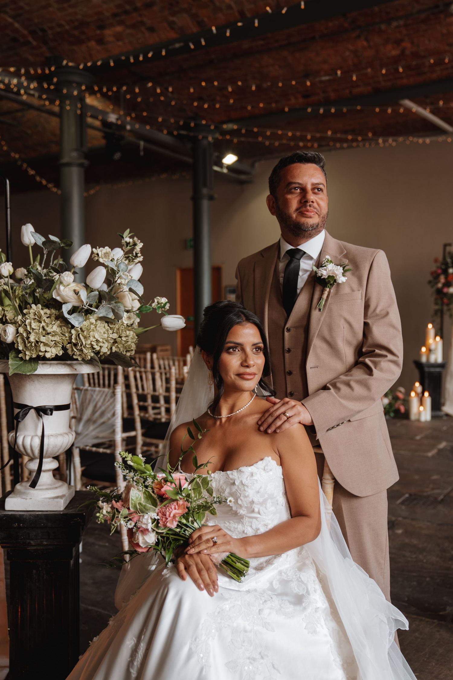 Bride in white gown holds bouquet while groom in tan suit stands behind her in elegant venue.