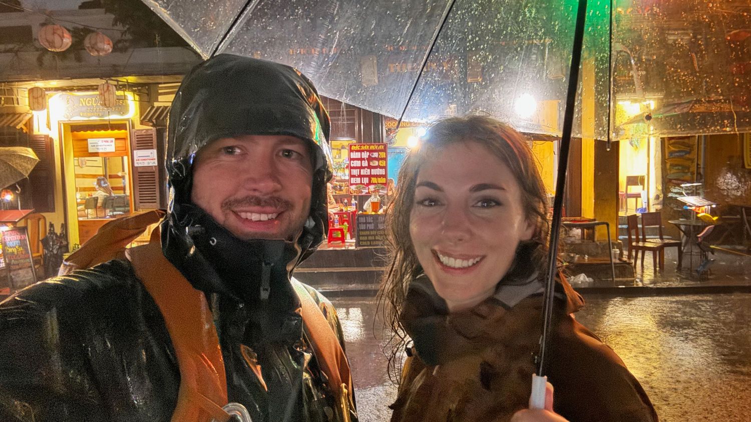 Kristen and Evan together under an umbrella on an extremely rainy, torrential night outside an illuminated storefront in Hoi An, Vietnam
