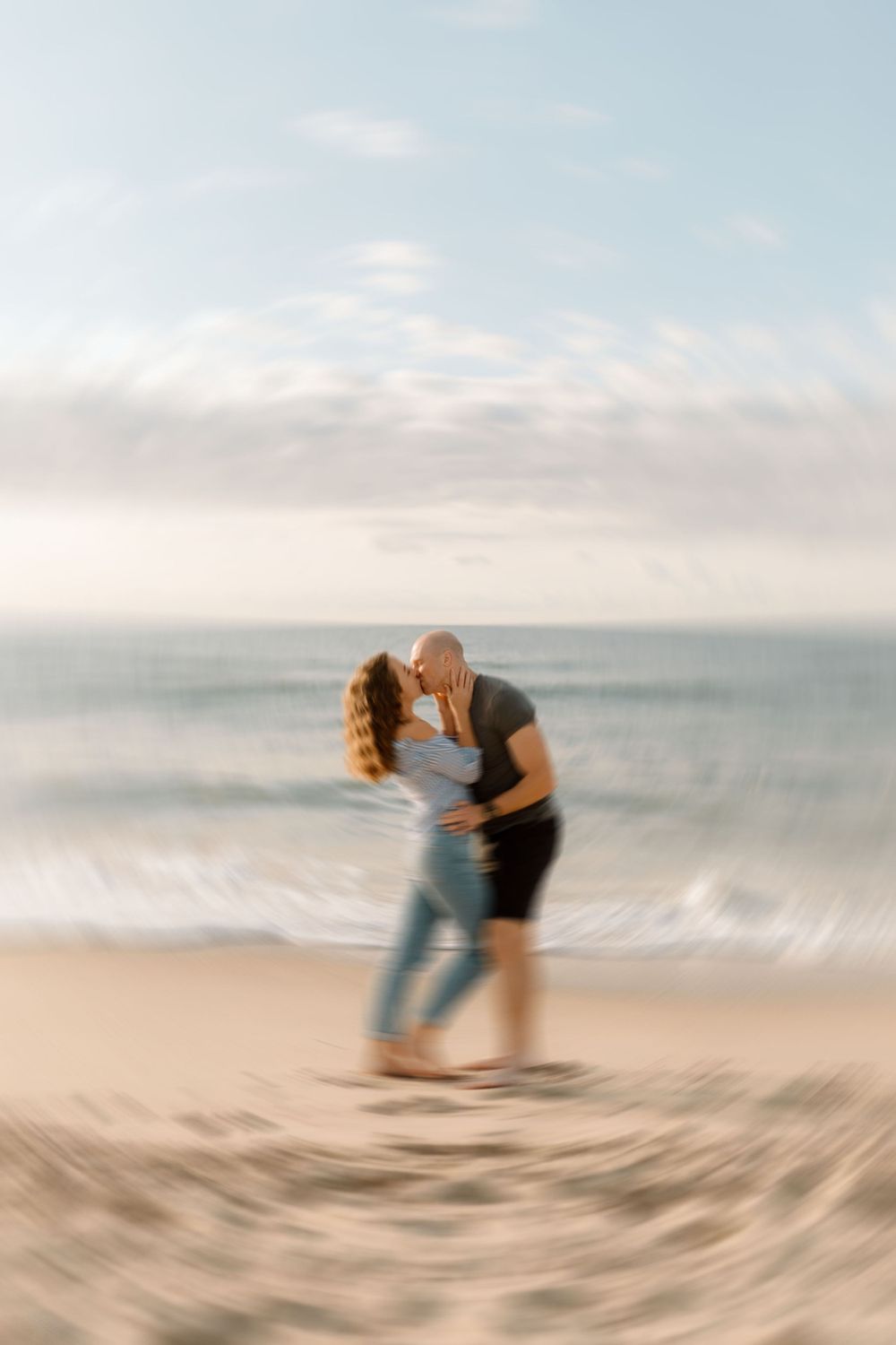 A blurred vignette surrounds a focused view of a couple kissing with the beach in the background, creating a dreamy, intimate atmosphere.