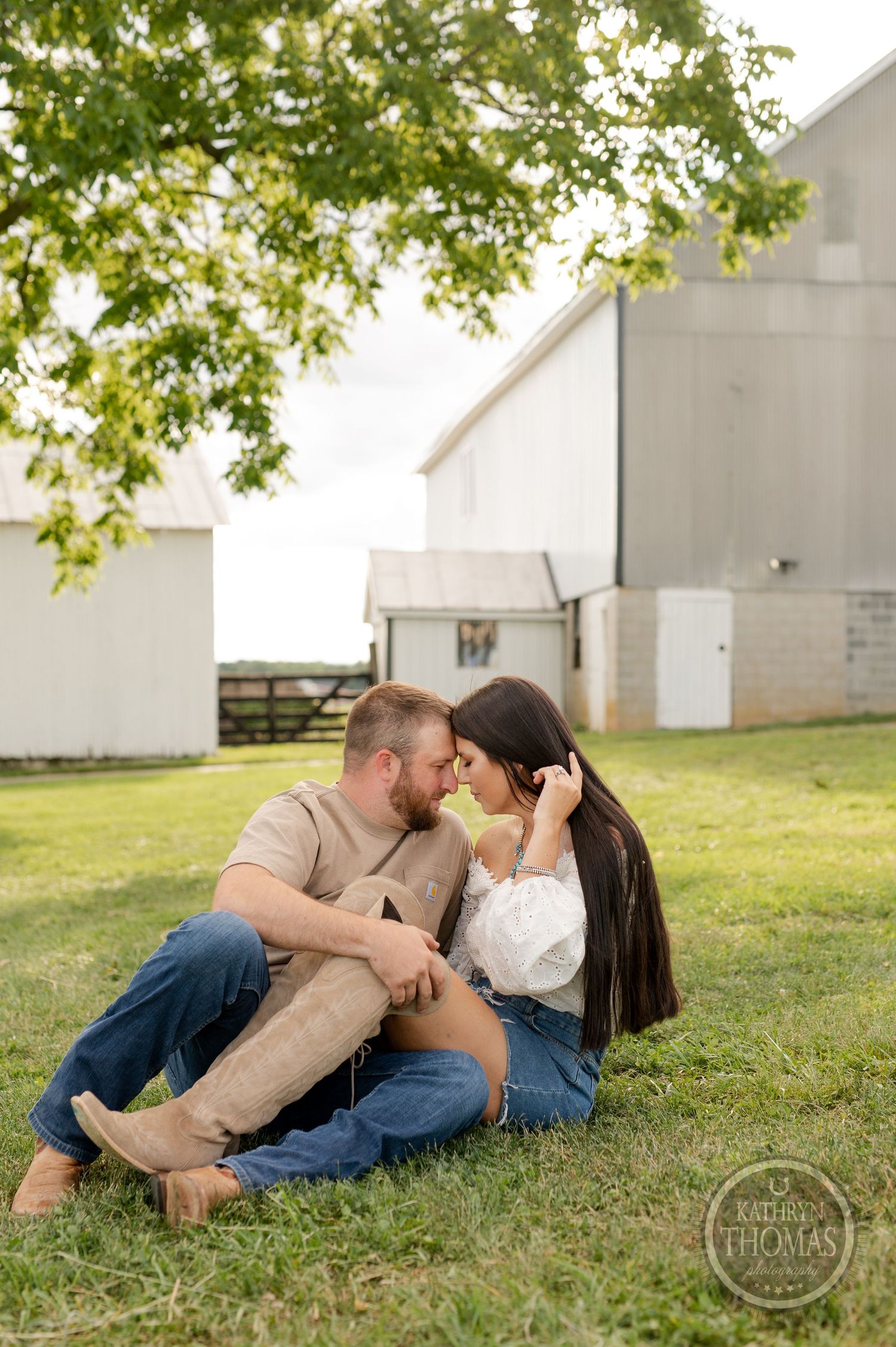 Intimate couple sitting close on grass with rustic barn setting in warm evening light.
