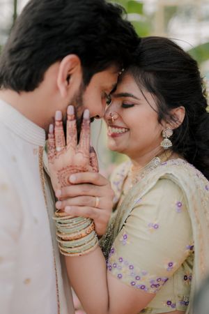 Indian couple in traditional attire sharing a romantic moment, bride adorned with mehndi, gold jewelry, and pastel lehenga.