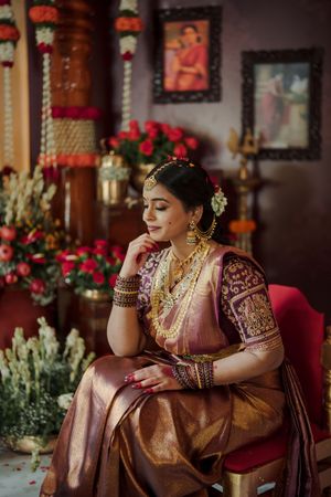 Traditional Indian bride in burgundy lehenga poses elegantly on red velvet sofa with floral decor in background.