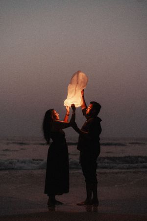 Couple releasing a glowing sky lantern on a beach at dusk, standing silhouetted against the ocean.