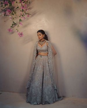 A model in an elegant silver-gray lehenga with floral embroidery poses in front of a pink floral backdrop.
