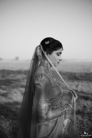 Black and white portrait of a bride in traditional Indian bridal attire with veil and jewelry, standing in an open field.