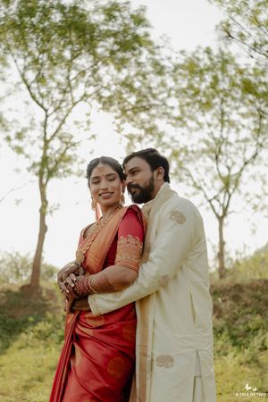 Indian bride in red saree and groom in cream sherwani embrace outdoors during traditional wedding photoshoot.