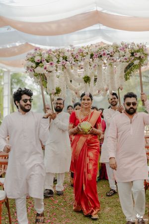 Bride in red saree walks under floral canopy held by groomsmen in white kurtas at outdoor South Indian wedding ceremony.
