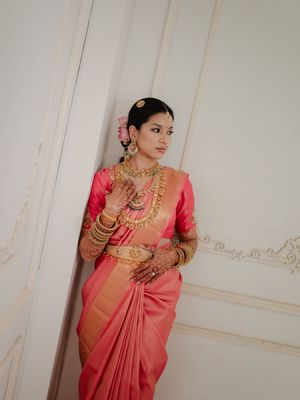 Traditional South Indian bride wearing a coral pink silk saree with ornate gold jewelry in an elegant indoor setting.