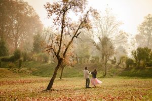 Couple holding hands in an autumn park surrounded by fallen leaves and misty trees in warm golden light.